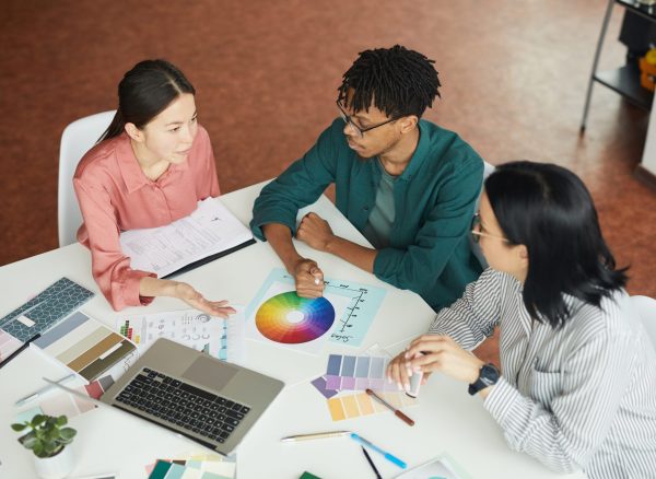 Multiethnic group of graphic designers sitting at the table with colored palette and choosing the color during their work at office